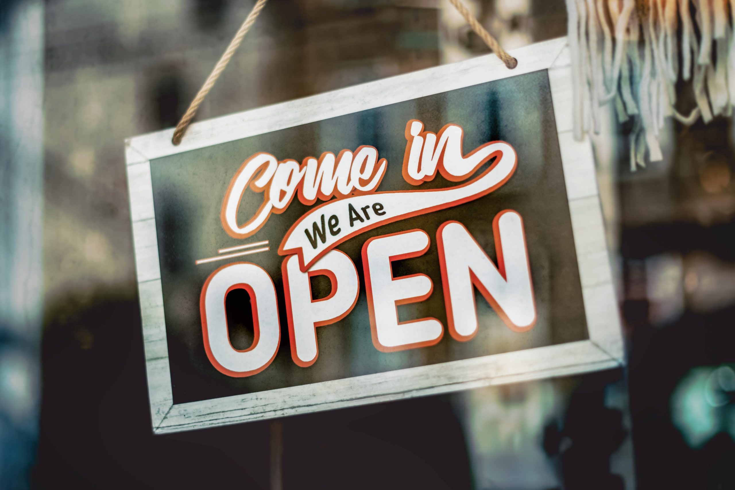 A welcoming 'Come in, We Are Open' sign hanging on a glass window of an urban shop.